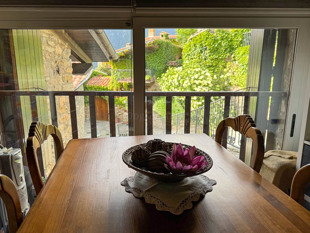 Menjador de Cal Marinall, amb una taula de fusta i cadires, al costat d'una finestra amb vistes a un carrer amb vegetació